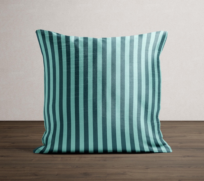 May include: Square throw pillow with a teal and dark teal vertical stripe pattern. The pillow is made of a soft fabric and is placed on a dark wood surface. The pillow is a decorative item for a bed or sofa.
