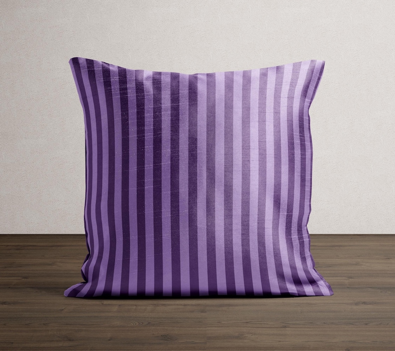 May include: A square decorative pillow with vertical stripes in shades of purple. The pillow is resting on a dark wooden surface. The stripes alternate between light and dark purple.