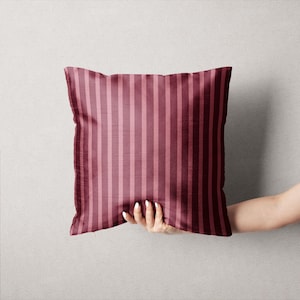 May include: A square decorative pillow with a vertical striped pattern in shades of burgundy and rose. The pillow is held by a hand against a neutral background. The pillow's design is simple and elegant.