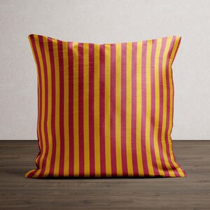 May include: A square decorative pillow with a vertical striped pattern. The stripes alternate between a deep red and a golden yellow. The pillow is placed on a dark wood surface.