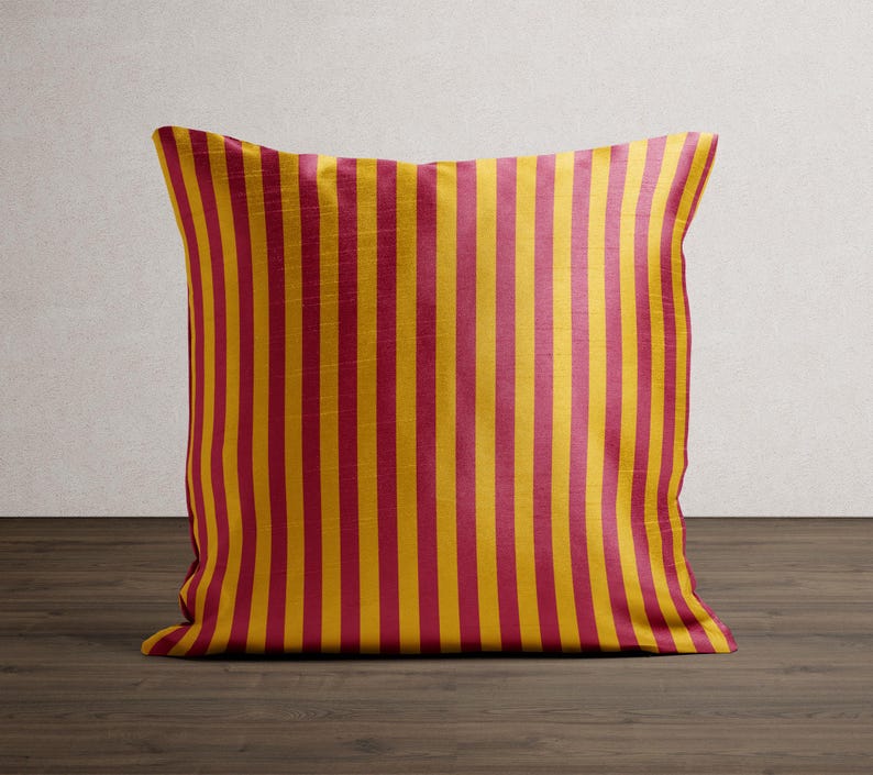 May include: A square decorative pillow with a vertical striped pattern. The stripes alternate between a deep red and a golden yellow. The pillow is placed on a dark wood surface.