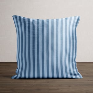 May include: A square pillow with a blue and light blue vertical stripe pattern. The pillow is resting on a dark brown wooden surface, with a neutral-colored wall in the background. The pillow is a decorative home accent.