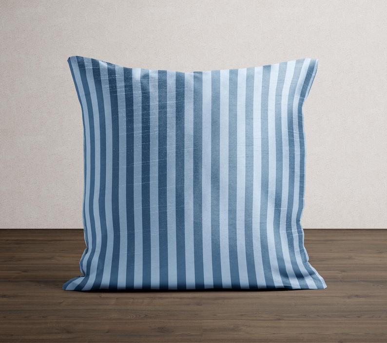 May include: A square pillow with a blue and light blue vertical stripe pattern. The pillow is resting on a dark brown wooden surface, with a neutral-colored wall in the background. The pillow is a decorative home accent.