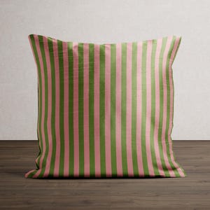 May include: A square decorative pillow with a vertical striped pattern. The stripes alternate between a muted green and a soft pink. The pillow is placed on a dark wood surface, with a neutral background.