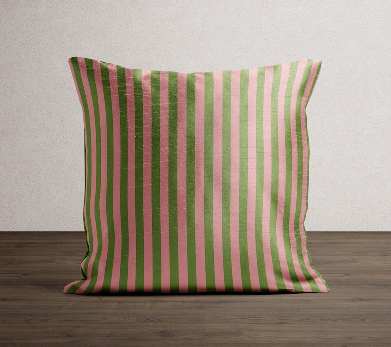 May include: A square decorative pillow with a vertical striped pattern. The stripes alternate between a muted green and a soft pink. The pillow is placed on a dark wood surface, with a neutral background.