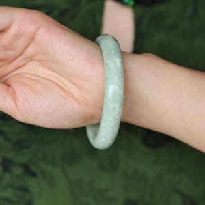 Pode incluir: Um bracelete liso e arredondado feito de pedra de jade verde pálido. A superfície do bracelete apresenta um acabamento polido com sutis inclusões brancas e nubladas por todo o material verde claro, criando uma forma circular sólida.