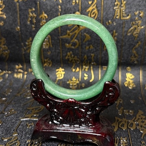 Può includere: Un bracciale rigido circolare in giada verde, liscio e dall'aspetto traslucido. Il bracciale è esposto su un supporto in legno scuro e levigato, caratterizzato da intricati dettagli intagliati lungo la base e i bracci di supporto curvi.