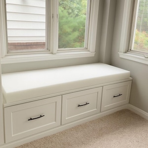 May include: A built-in window seat with three white drawers and a thick, white cushion. The drawers have black handles. The window seat is positioned beneath a large window with a view of greenery.