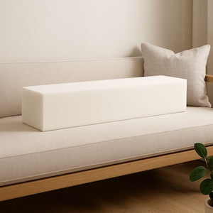 May include: An off-white rectangular foam bolster cushion sits on a beige sofa. The cushion is a solid block shape. A beige throw cushion is in the background. The sofa has a wooden frame. A houseplant is in the lower right corner.