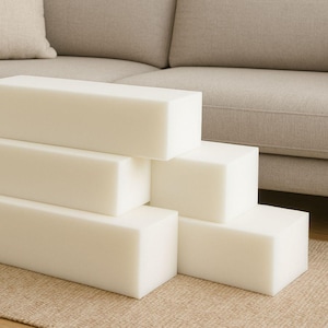 May include: Several off-white foam blocks, rectangular in shape, are stacked in a stepped arrangement. The blocks are in front of a beige sofa, resting on a light brown rug. Each block is approximately 30 cm long.