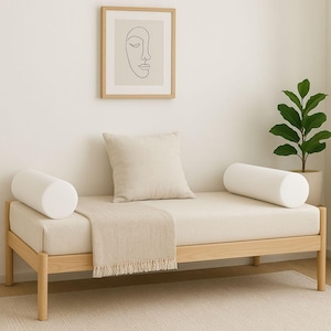 May include: A light wood daybed with a cream cushion, two white bolster pillows, and a square pillow. A beige throw blanket with fringe is draped over the bed. A framed line art print and a potted plant are in the background.