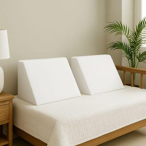 May include: Two white foam bed wedges are placed on a bed covered with a white quilted bedspread. The wedges are triangular, designed to elevate the head and upper body. The bed frame is made of wood, and a lamp and plant are visible in the background.