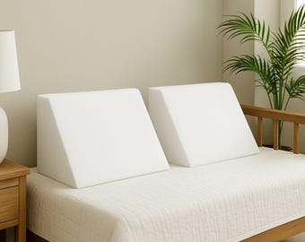 May include: Two white foam bed wedges are placed on a bed covered with a white quilted bedspread. The wedges are triangular, designed to elevate the head and upper body. The bed frame is made of wood, and a lamp and plant are visible in the background.