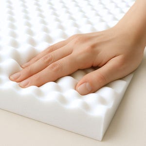 May include: A close-up of a white foam mattress topper with a textured surface. A hand is gently pressing into the foam, showing its softness and flexibility. The foam has a wave-like pattern.