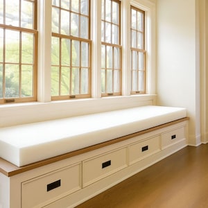 May include: A built-in window seat with a white cushion and drawers. The seat is positioned beneath a row of wooden-framed windows. The drawers have black rectangular handles. The room has hardwood flooring.