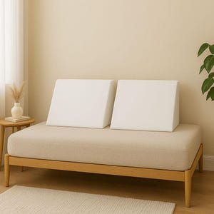 May include: A beige upholstered daybed with two white, wedge-shaped pillows. The bed frame is made of light-colored wood. A small side table with a vase and dried flowers is to the left. A rug is in the foreground.