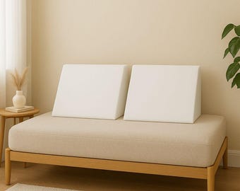 May include: A beige upholstered daybed with two white, wedge-shaped pillows. The bed frame is made of light-colored wood. A small side table with a vase and dried flowers is to the left. A rug is in the foreground.