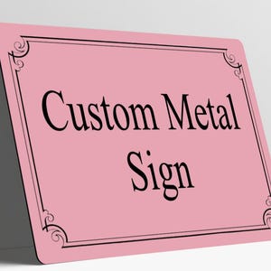 May include: A rectangular pink metal sign featuring a black border with decorative corner flourishes. The text Custom Metal Sign is printed in the center in a black serif font. The sign has rounded corners and a smooth, flat surface.