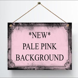 May include: A rectangular sign with a pale pink background and black text that reads "*NEW* PALE PINK BACKGROUND". The sign has a black border with decorative corner accents and is suspended by a brown rope.