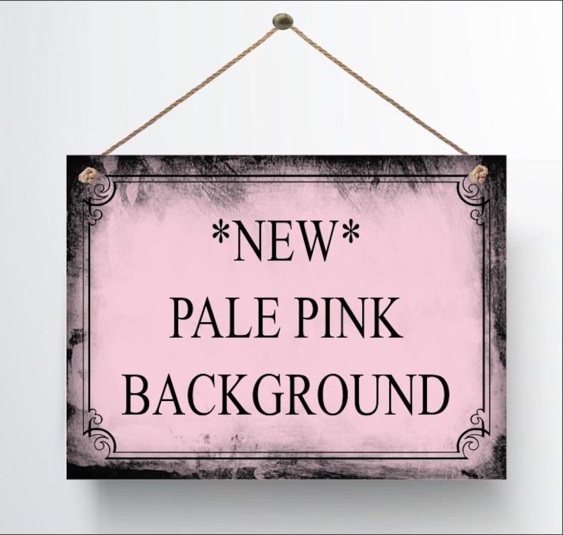 May include: A rectangular sign with a pale pink background and black text that reads "*NEW* PALE PINK BACKGROUND". The sign has a black border with decorative corner accents and is suspended by a brown rope.