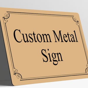May include: A rectangular tan metal sign with rounded corners featuring a black border with decorative scrollwork in each corner. The text "Custom Metal Sign" is printed in a black serif font in the center of the sign.