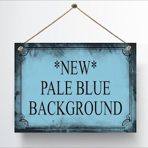 May include: A rectangular sign with a pale blue background and black text that reads "*NEW* PALE BLUE BACKGROUND". The sign has a dark border with decorative corner accents and is suspended by a brown rope.