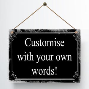 May include: A rectangular black sign with white text that reads "Customise with your own words!" The sign has a decorative white border and is suspended by a brown rope. The sign is hanging against a white background.