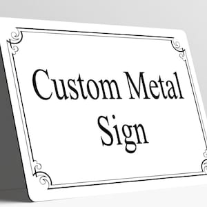 May include: A rectangular white metal sign with rounded corners, featuring a black decorative border with scrollwork at each corner. The text Custom Metal Sign is printed in the center in a black serif font, oriented horizontally.