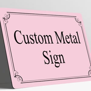 May include: A rectangular light pink metal sign with rounded corners. The sign features a black double-line border with decorative scrollwork at each corner. The text Custom Metal Sign is printed in the center in a black serif font.