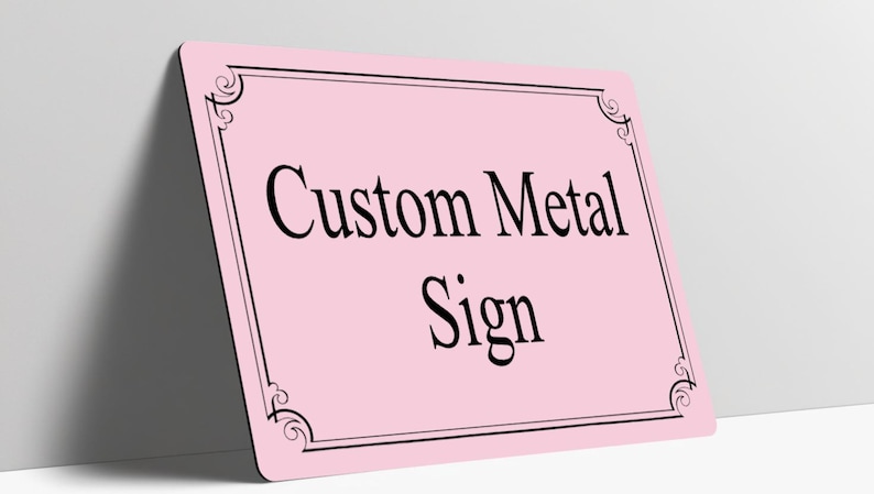 May include: A rectangular light pink metal sign with rounded corners. The sign features a black double-line border with decorative scrollwork at each corner. The text Custom Metal Sign is printed in the center in a black serif font.