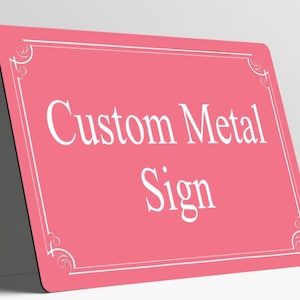 May include: A rectangular pink metal sign with rounded corners featuring a white border with decorative corner flourishes. The text Custom Metal Sign is printed in the center in a white serif font. The sign is positioned at an angle.