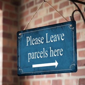 May include: A rectangular blue sign with white text that reads "Please Leave parcels here" and a white arrow pointing right. The sign is hanging from a black metal bracket on a brick wall.