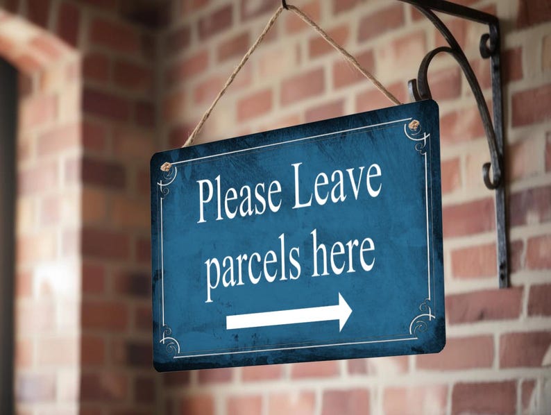 May include: A rectangular blue sign with white text that reads "Please Leave parcels here" and a white arrow pointing right. The sign is hanging from a black metal bracket on a brick wall.