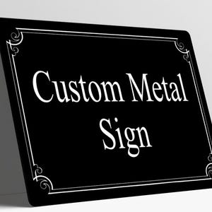 May include: A rectangular black metal sign with rounded corners featuring the white text Custom Metal Sign in a serif font. The sign has a thin white border with decorative scrollwork accents in each of the four corners.