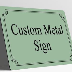 May include: A rectangular sage green metal sign featuring a black border with decorative corner flourishes. The text Custom Metal Sign is printed in the center in a black serif font. The sign has rounded corners and a smooth finish.