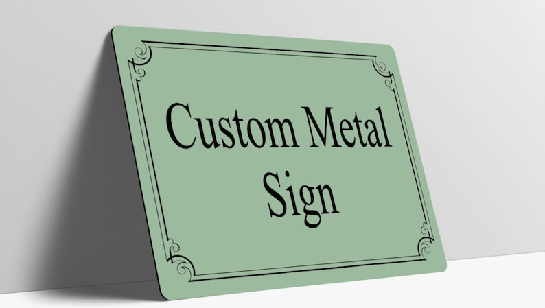 May include: A rectangular sage green metal sign featuring a black border with decorative corner flourishes. The text Custom Metal Sign is printed in the center in a black serif font. The sign has rounded corners and a smooth finish.
