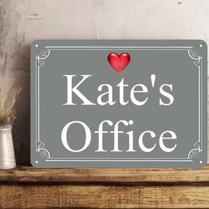 May include: A rectangular gray metal sign with white decorative borders and the text "Kate's Office" in white lettering. A small, glossy red heart icon is positioned above the text. The sign features four small holes in the corners.