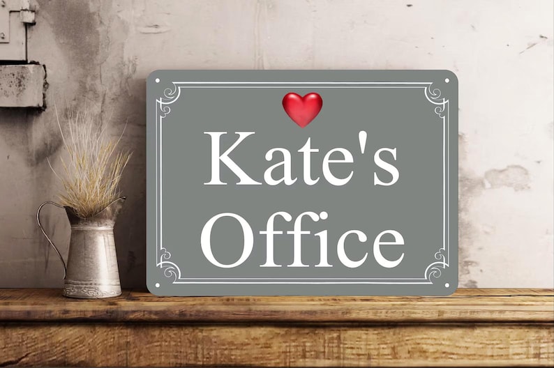 May include: A rectangular gray metal sign with white decorative borders and the text "Kate's Office" in white lettering. A small, glossy red heart icon is positioned above the text. The sign features four small holes in the corners.