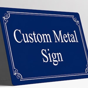 May include: A rectangular dark blue metal sign with rounded corners. The sign features a white decorative border and the white text Custom Metal Sign centered on the surface. The sign is positioned at an angle against a plain white wall.
