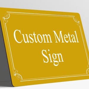May include: A rectangular gold metal sign with rounded corners featuring a white border with decorative corner flourishes. The text Custom Metal Sign is printed in white in the center of the sign in a serif font.