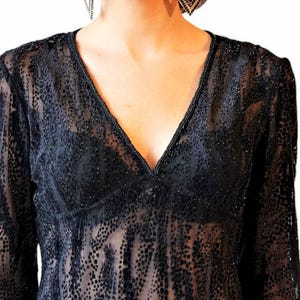 May include: Black sheer long-sleeve top with a V-neckline. The top is embellished with small, dark dots and a beaded trim along the neckline. The model is wearing dangling earrings.