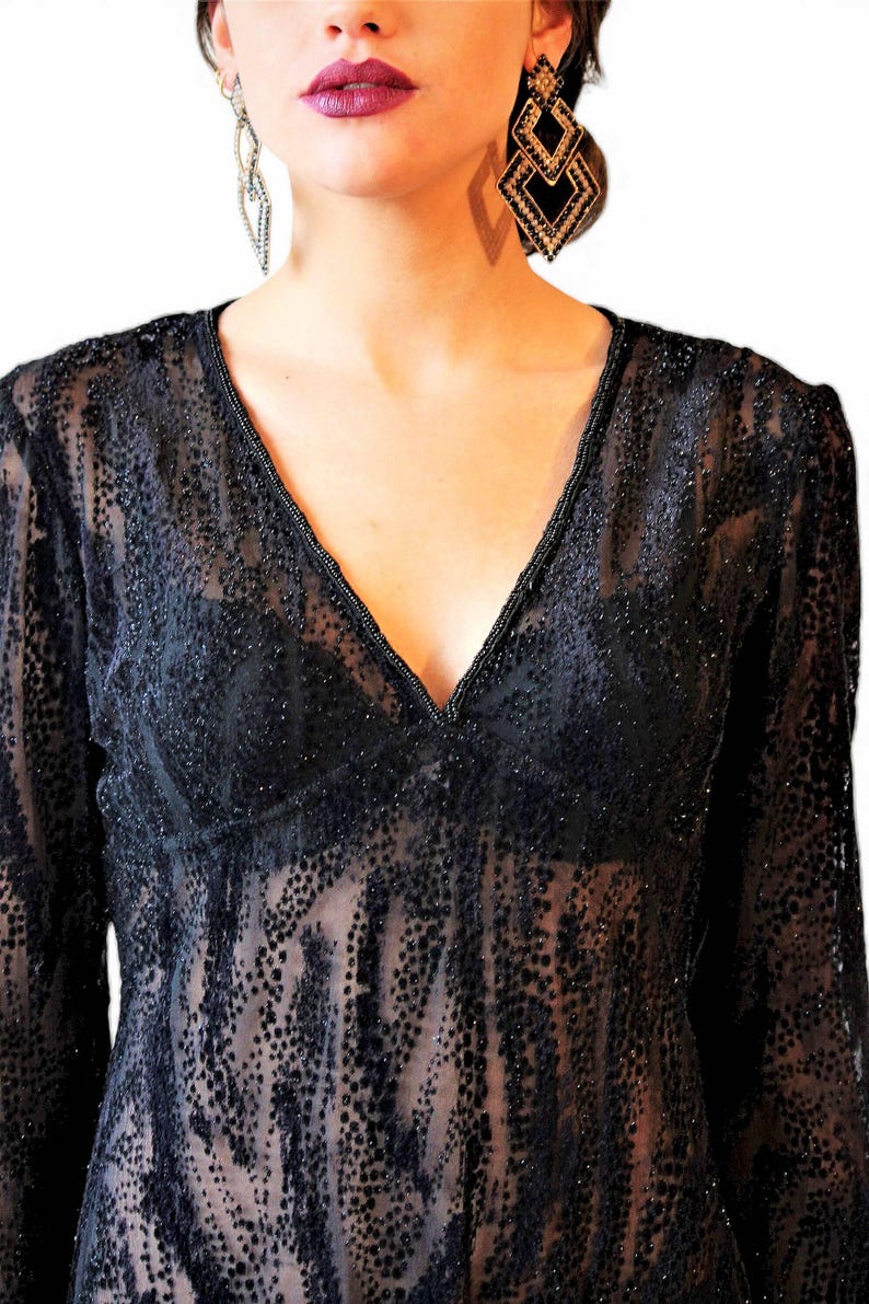 May include: Black sheer long-sleeve top with a V-neckline. The top is embellished with small, dark dots and a beaded trim along the neckline. The model is wearing dangling earrings.