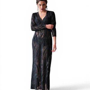 May include: A full-length black lace dress with long sleeves and a V-neck. The dress has a flowing, floor-length skirt. The model is wearing black and gold heels. The dress is made of a sheer, patterned fabric.