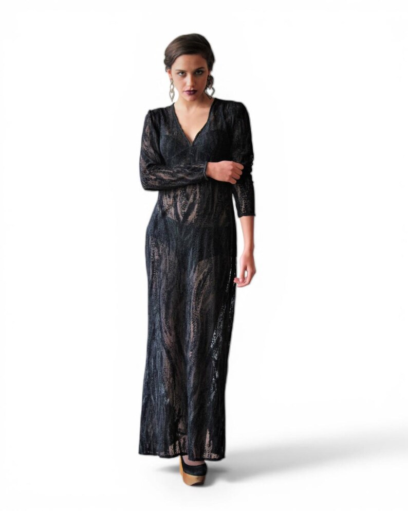 May include: A full-length black lace dress with long sleeves and a V-neck. The dress has a flowing, floor-length skirt. The model is wearing black and gold heels. The dress is made of a sheer, patterned fabric.
