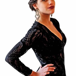 May include: A black, long-sleeved dress with a plunging neckline. The dress is covered in sequins and has a textured pattern. The model is wearing geometric drop earrings.
