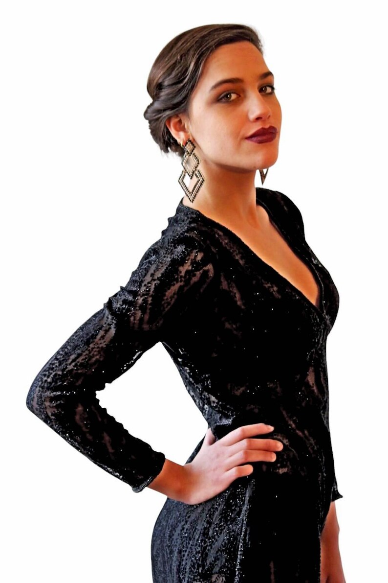 May include: A black, long-sleeved dress with a plunging neckline. The dress is covered in sequins and has a textured pattern. The model is wearing geometric drop earrings.