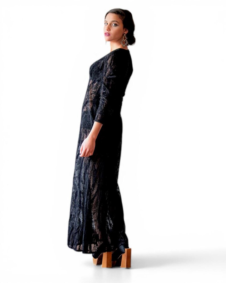 May include: A black lace maxi dress with long sleeves. The dress has a V-neckline and a flowing skirt. The model is wearing black platform shoes with wooden heels. The background is white.