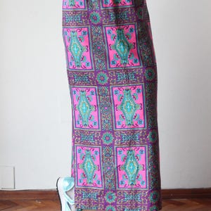 May include: A long, straight-cut skirt featuring a vibrant grid pattern of magenta and purple rectangles. Each section contains intricate teal, blue, and gold geometric motifs. The skirt has a solid purple waistband and is paired with metallic light blue sneakers.