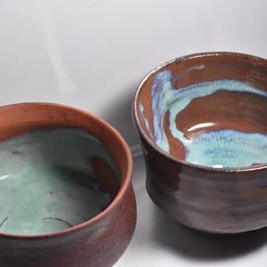 May include: Two ceramic bowls with a brown glaze and a light blue glaze inside. The bowls are handmade and have a rustic look.