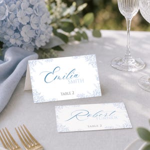 Puede incluir: Dos tarjetas de lugar blancas plegadas con un borde floral azul claro. Las tarjetas muestran los nombres Emilia Smith y Robert Turner en una fuente cursiva azul, con el texto TABLE 2 impreso en una fuente sans-serif bajo cada nombre.